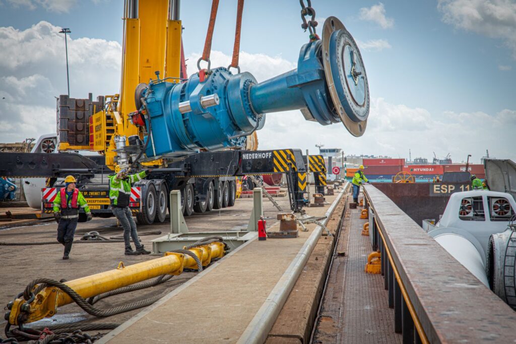 Twee werkers in veiligheidskleding sturen een groot blauw turbine-onderdeel aan dat in de lucht hangt aan de kabels van een hijskraan.