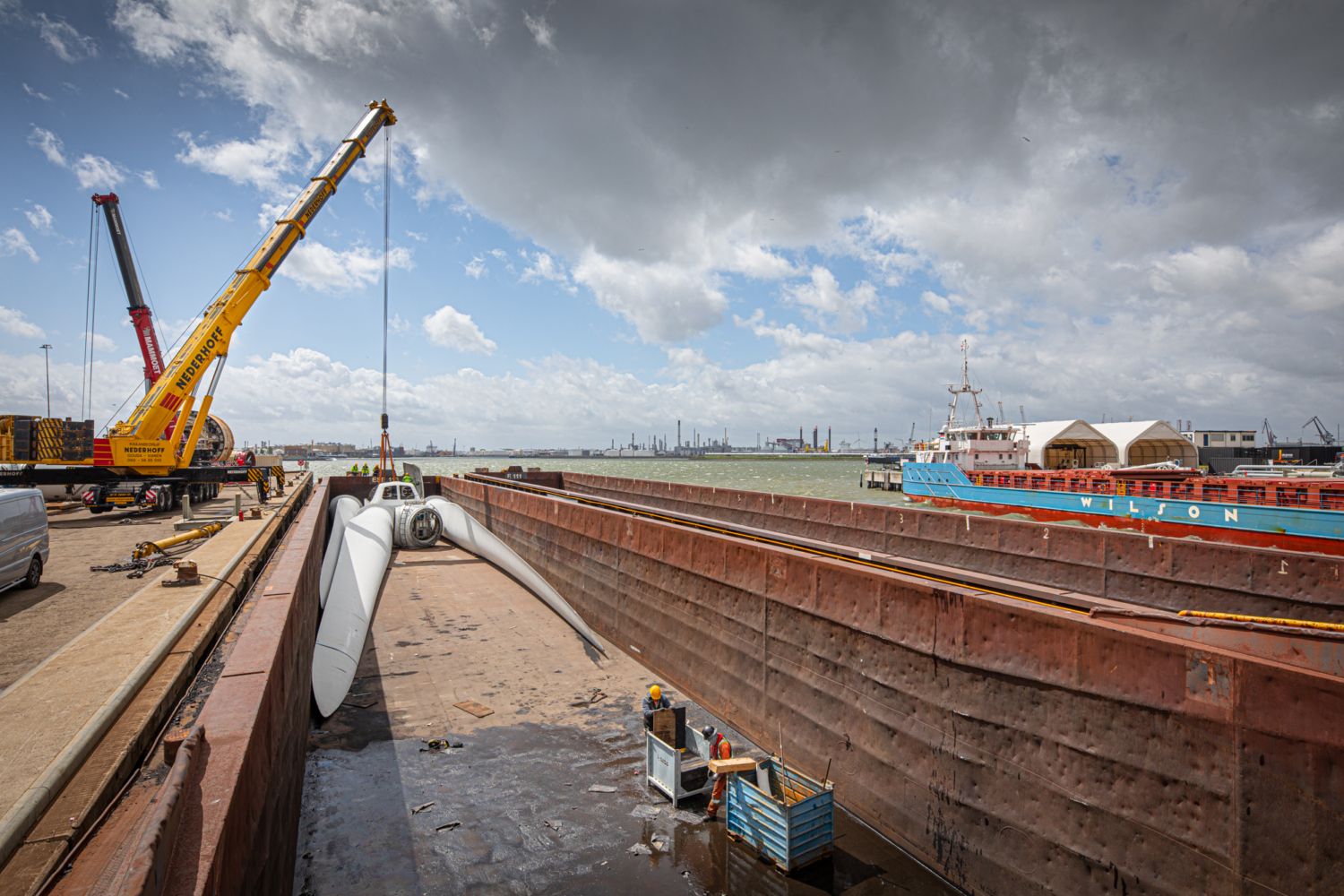 Een havengezicht overdag waar een grote hijskraan witte windturbinebladen in het laadruim van een groot vrachtschip plaatst.