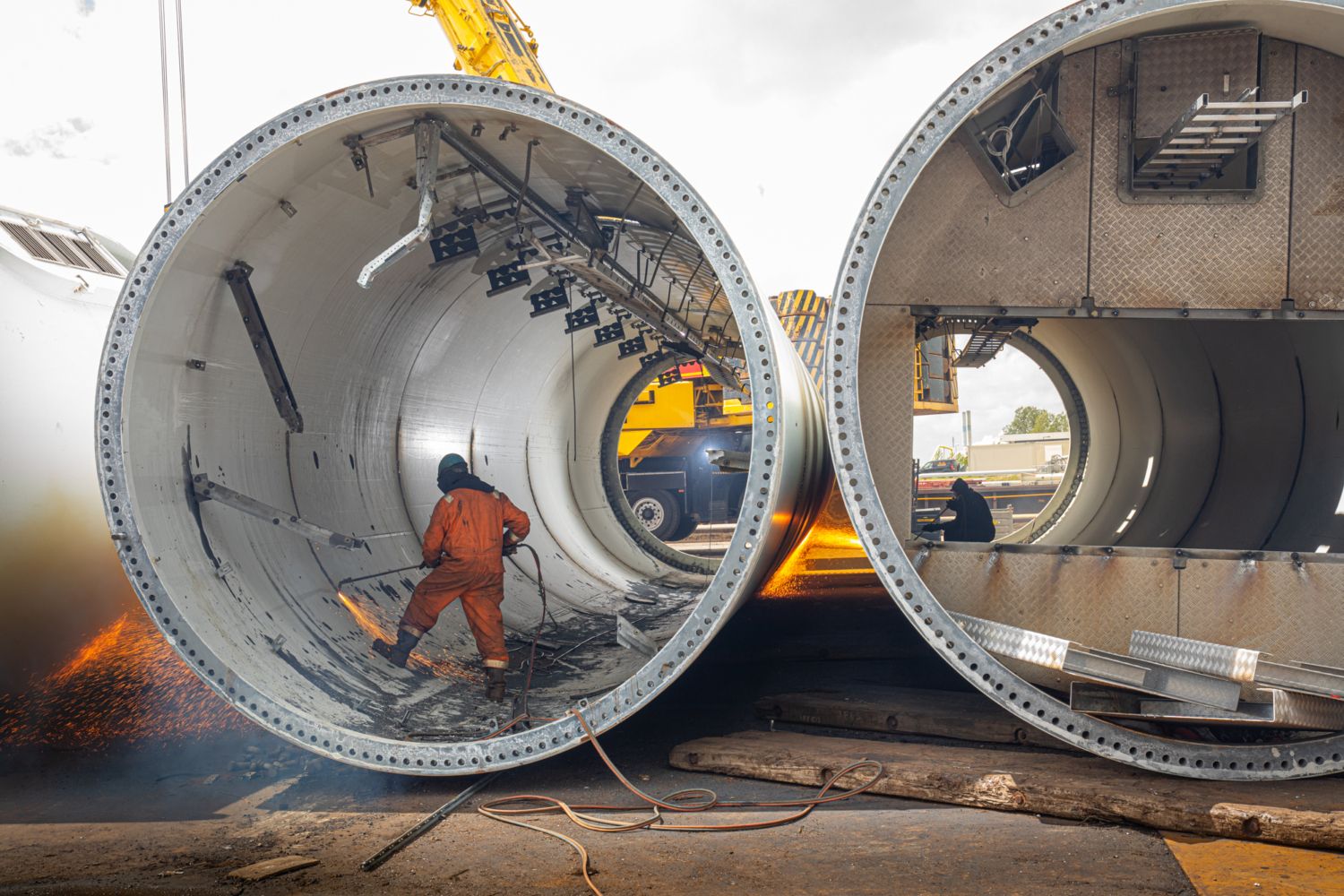 Een arbeider in een oranje pak gebruikt een snijbrander in een groot, cilindrisch metalen onderdeel van een windturbine, met wegschietende vonken.