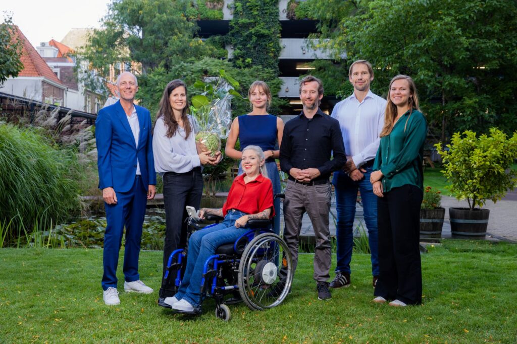 Groepsfoto van zes personen in een groene tuin, waaronder een rolstoelgebruiker